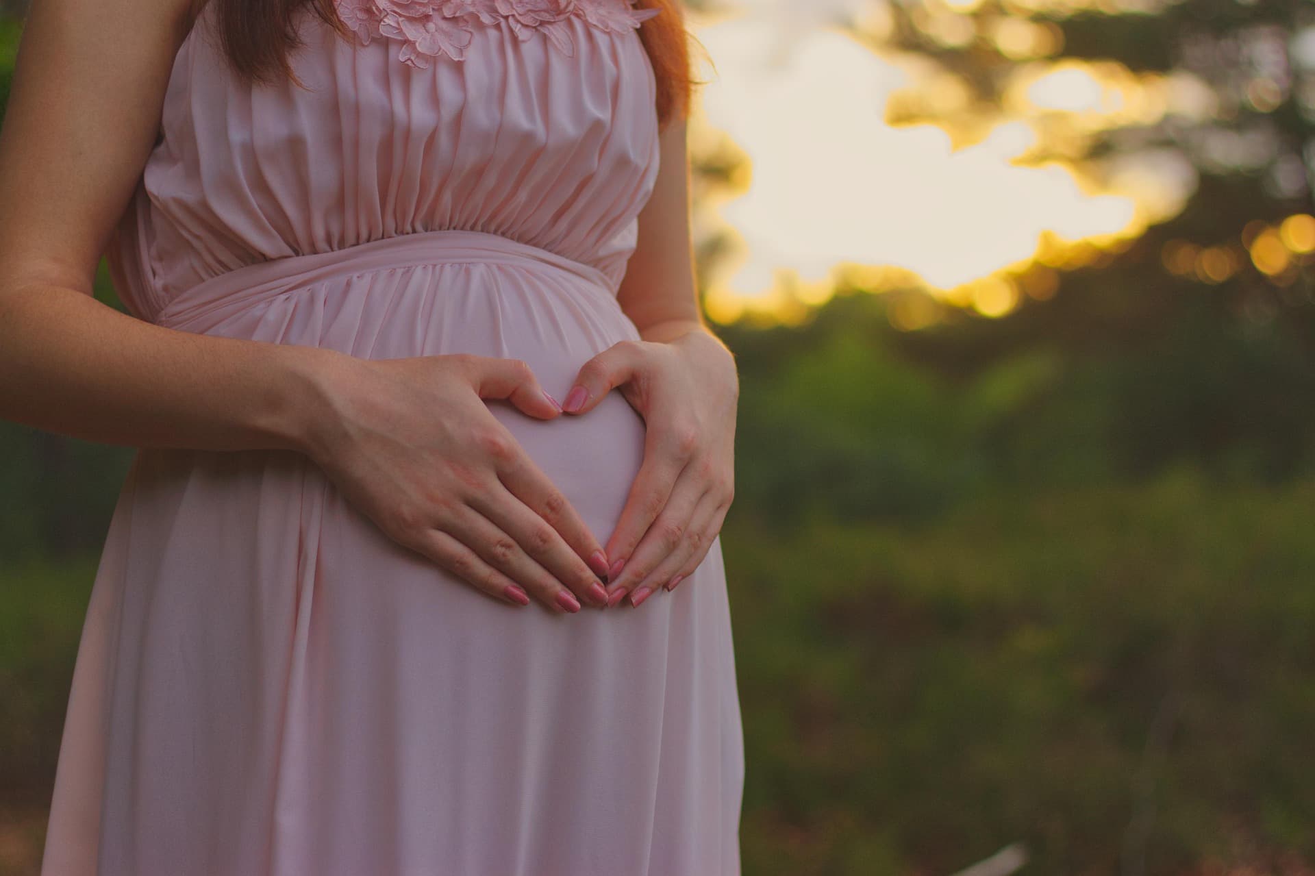 A pregnant woman in a pink dress has heart-shaped hands over her belly